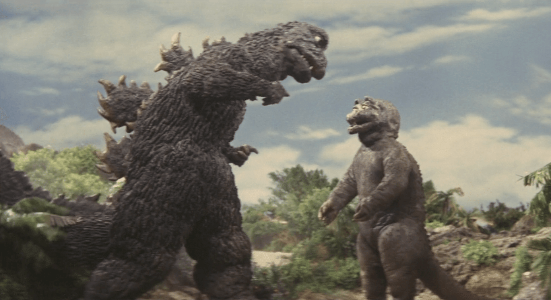 A Showa-era Godzilla facing the smaller, round-faced Minilla on a rocky island with trees and a blue sky.