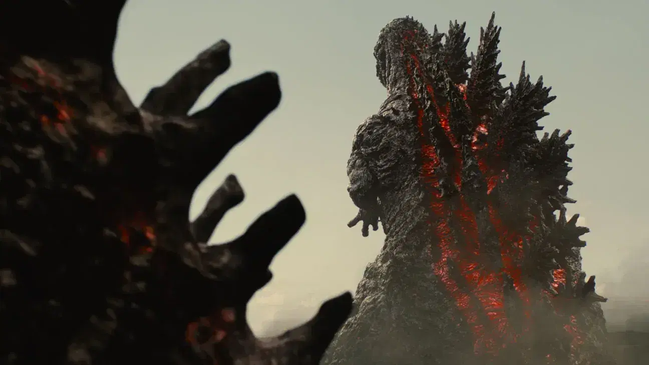 Shin Godzilla seen from behind in the distance, red light glowing through the seams of his dark ridged back, with a blurred dark hand in the foreground.