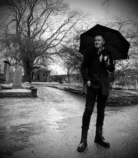 Man in a dark coat and boots stands in a graveyard holding an umbrella. Leafless trees and gravestones surround him, conveying a somber mood.