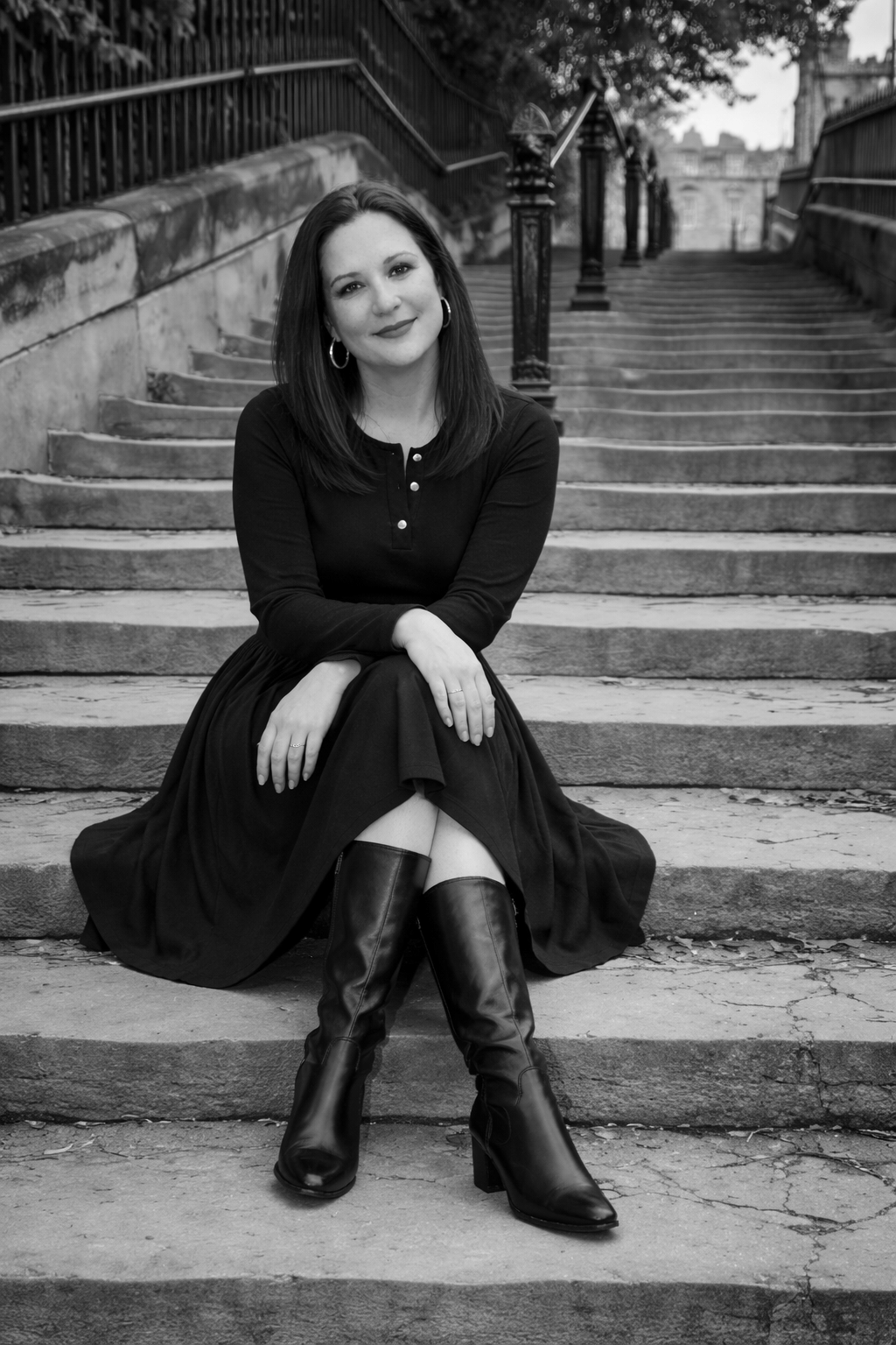 A woman in a black dress and boots sits on stone steps, hands clasped, smiling gently. The mood is serene and timeless, with a classic backdrop.