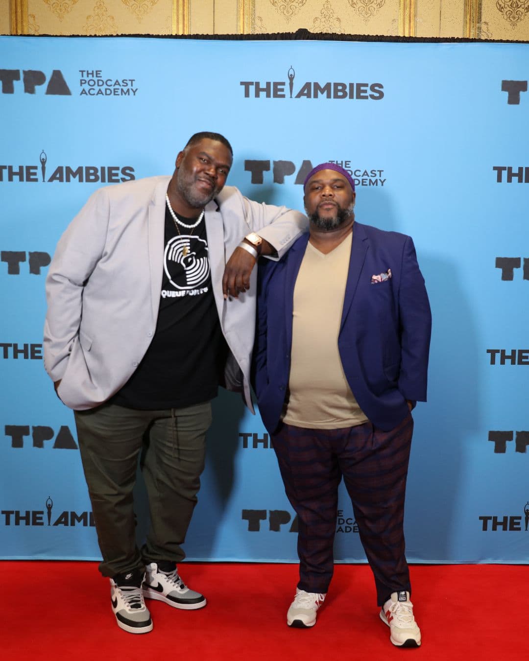 Two men stand side by side on a red carpet in front of a blue backdrop with "The Ambies" and "The Podcast Academy" logos.