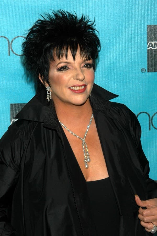 A woman with short, spiky black hair, wearing a black outfit and a sparkling necklace, smiles in front of a blue background with white and black text and logos.