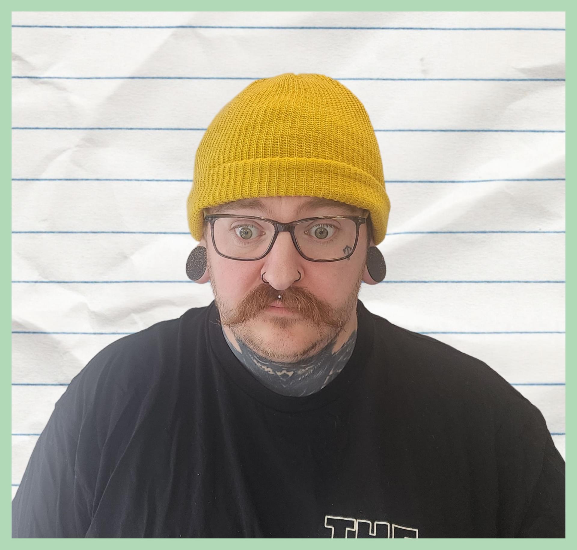 Carter in a mustard yellow beanie wearing glasses with a stylish moustache looking bewildered
